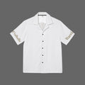 A white short-sleeve camp collar button-up shirt with black buttons, featuring 'Valhalla' printed in gold on the right sleeve and 'Warri' in gold on the left sleeve, displayed flat against a white background.